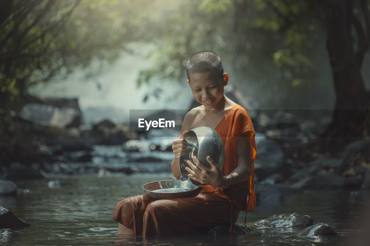 Smiling monk holding bowl and plate while sitting on rock amidst stream