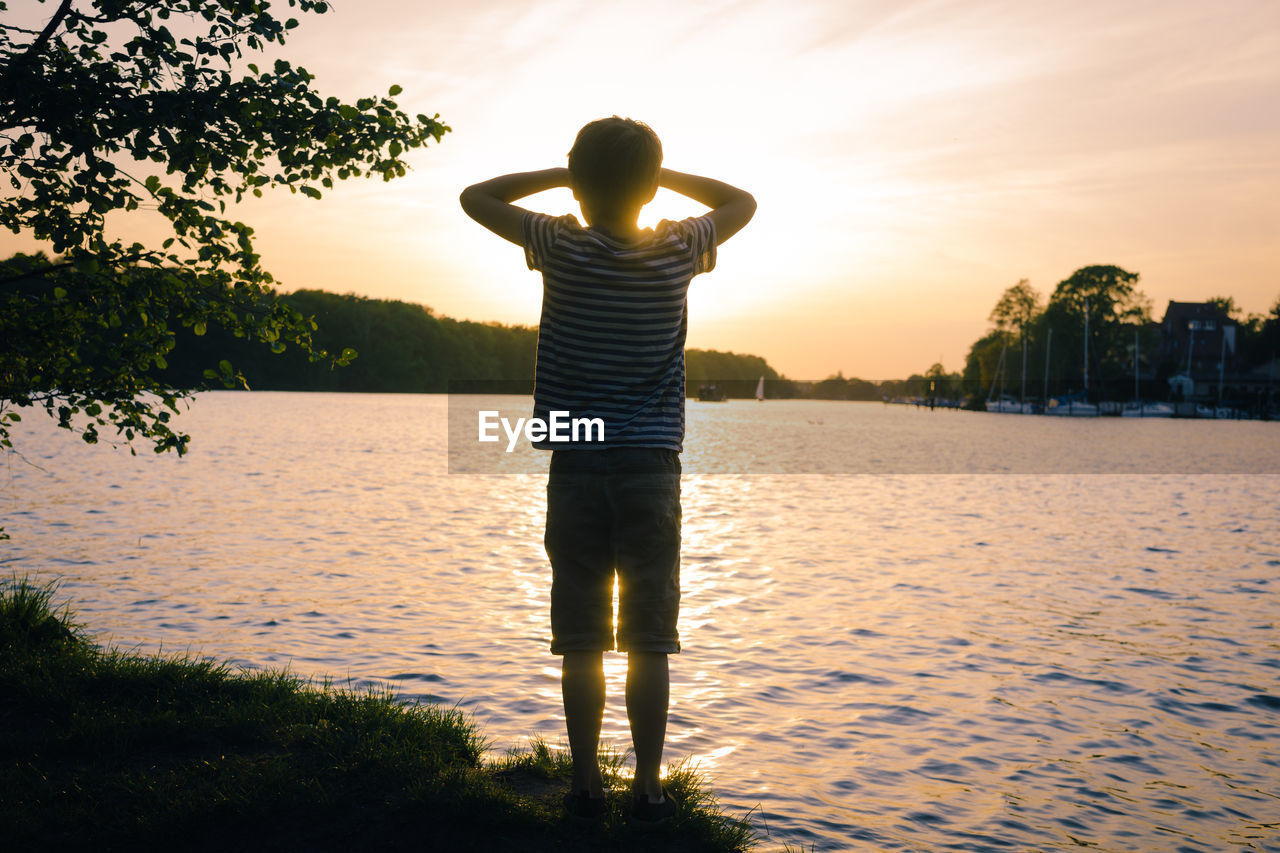 Rear view of boy standing by lake during sunset