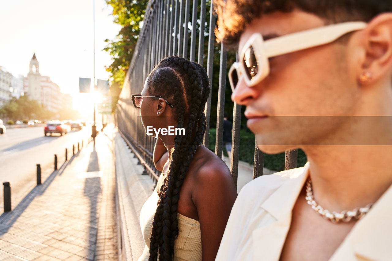 Stylish black woman with braided hair looking at road while leaning on park fence near boyfriend at sundown in city