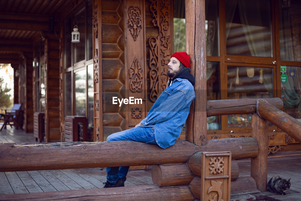 Portrait of a village man with a beard in a red hat stands at a wooden house in autumn