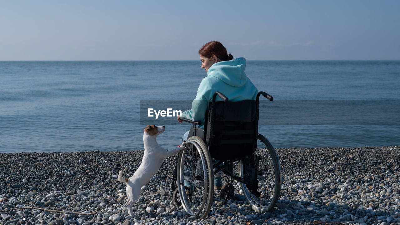 Caucasian woman in a hoodie in a wheelchair with a dog on the seashore.