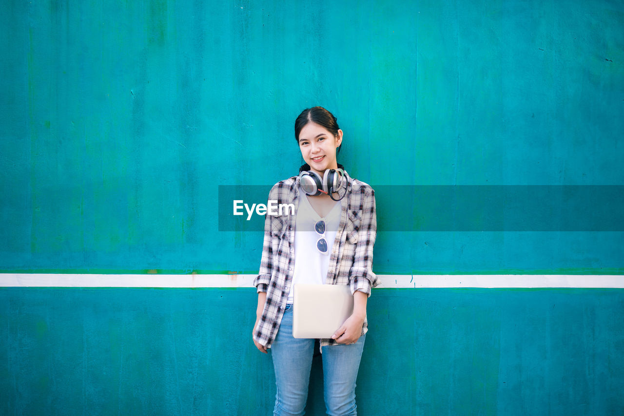 Young woman with laptop and headphones standing against blue wall