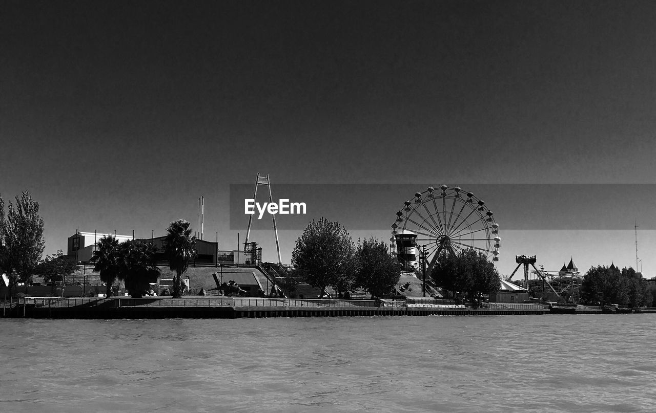 FERRIS WHEEL BY RIVER AGAINST BUILDINGS