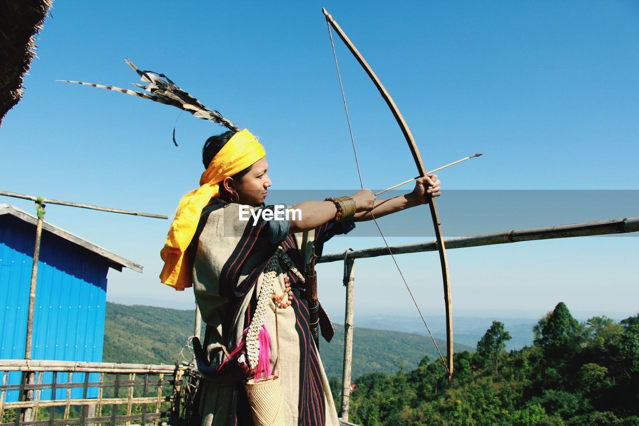 Man holding bow and arrow while standing against sky