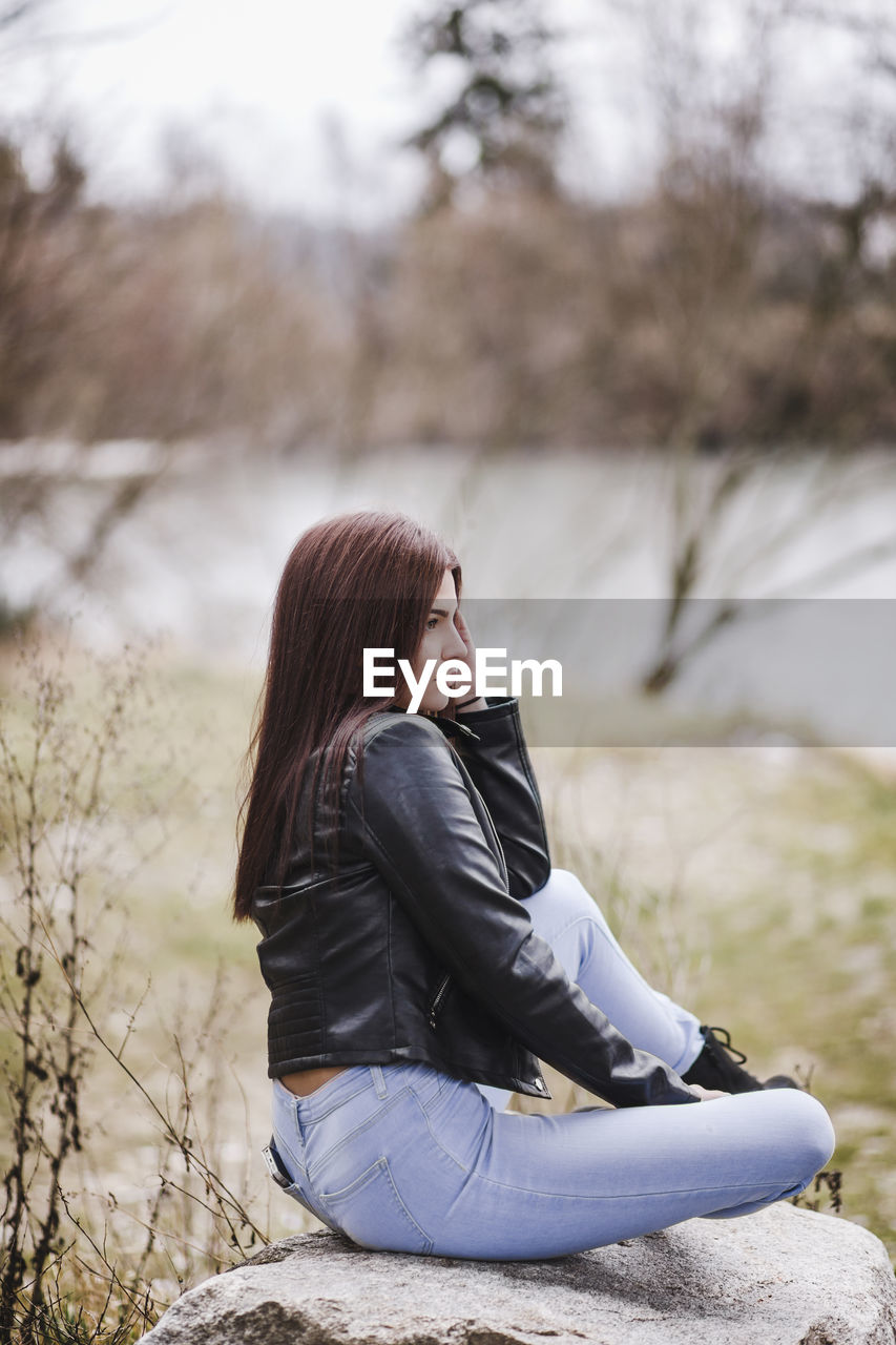 Side view of thoughtful teenage girl wearing leather jacket while sitting on rock