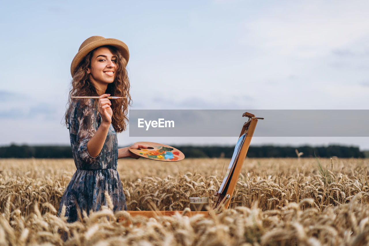 A young woman with curly hair and wearing a hat is painting in nature. 