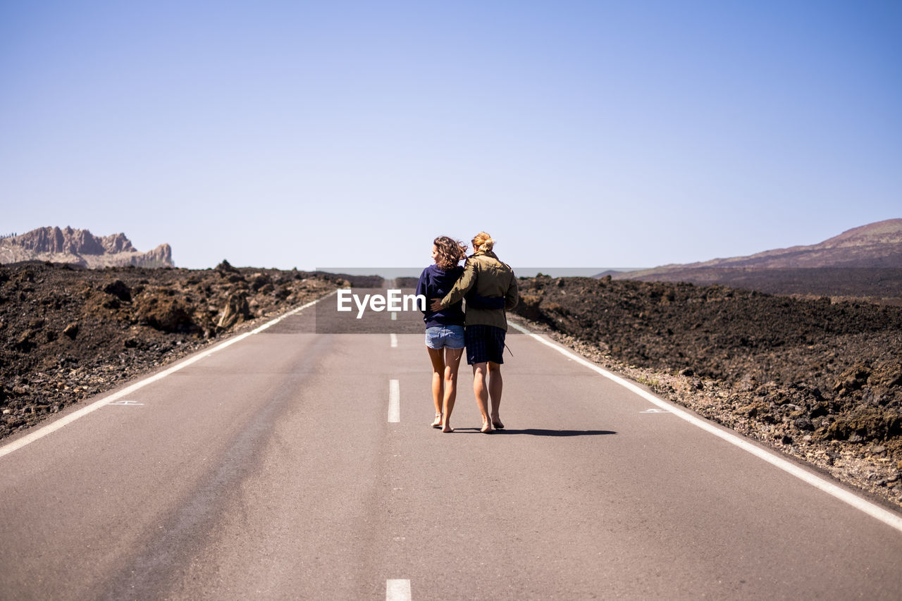 Rear view of young couple walking on road against sky during sunny day