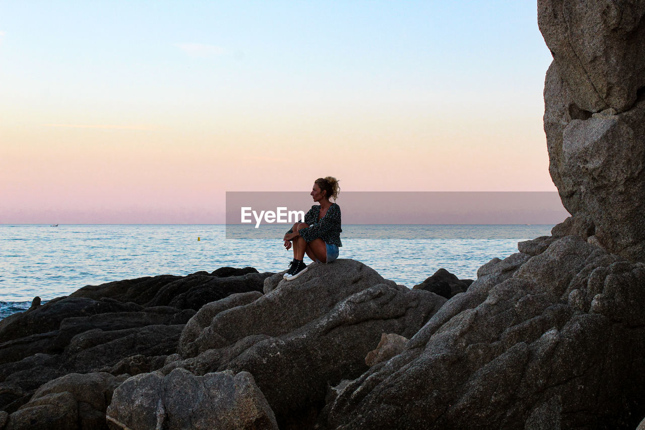 Woman sitting on rock by sea against sky during sunset