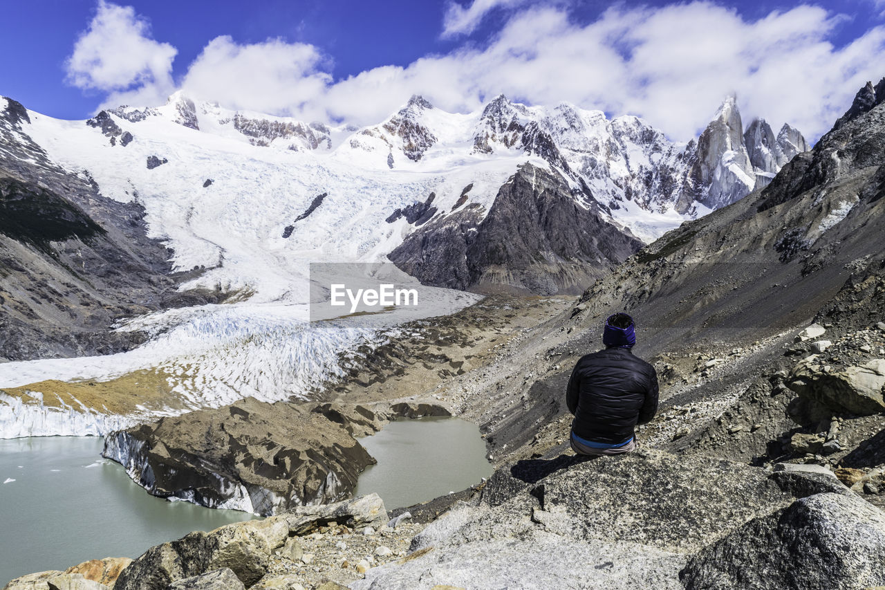 Rear view of person sitting on rock against snowcapped mountain range