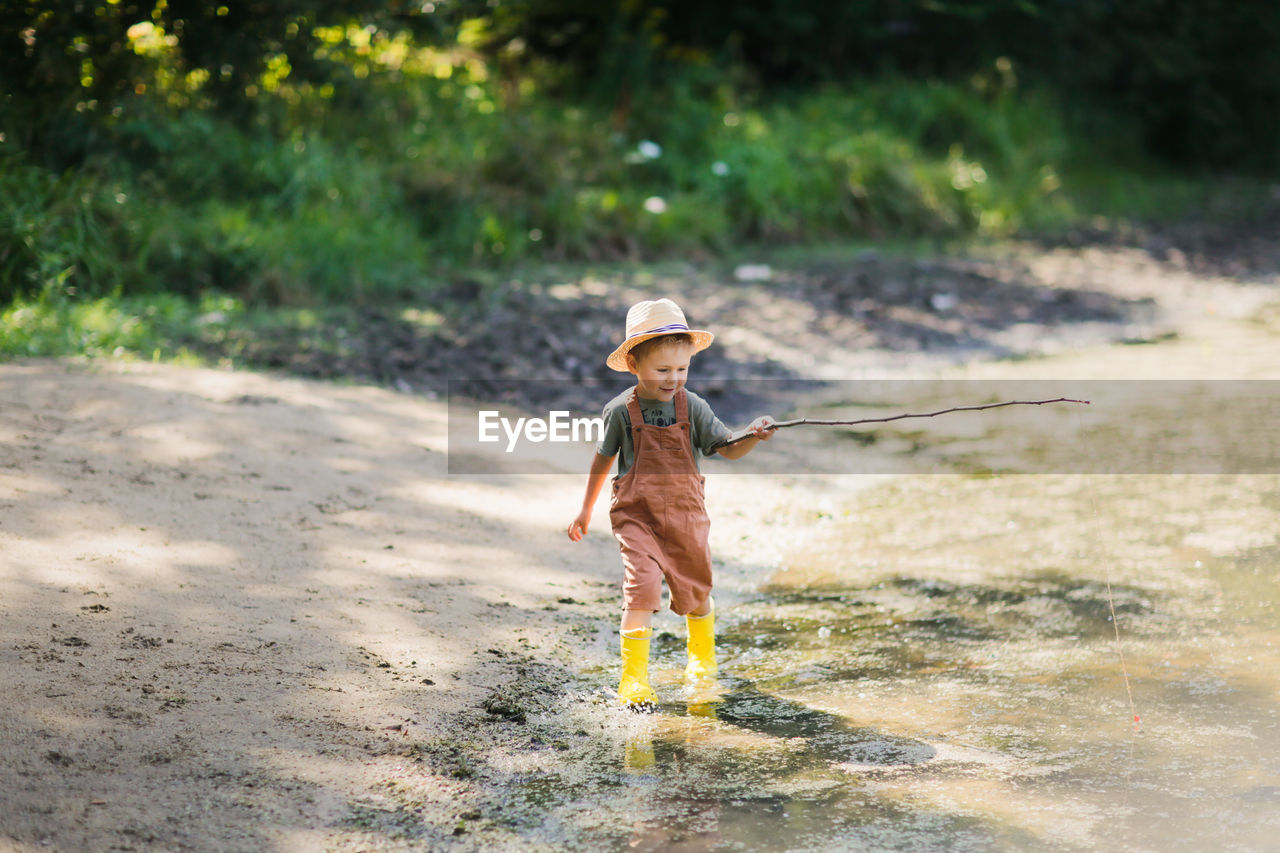 Preschooler boy in yellow rubber boots plays fishing, child fishes with stick  in countryside