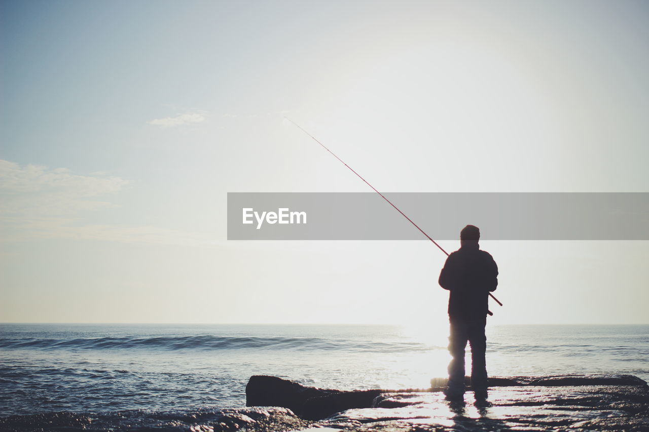 Rear view of man fishing in sea against sky on sunny day