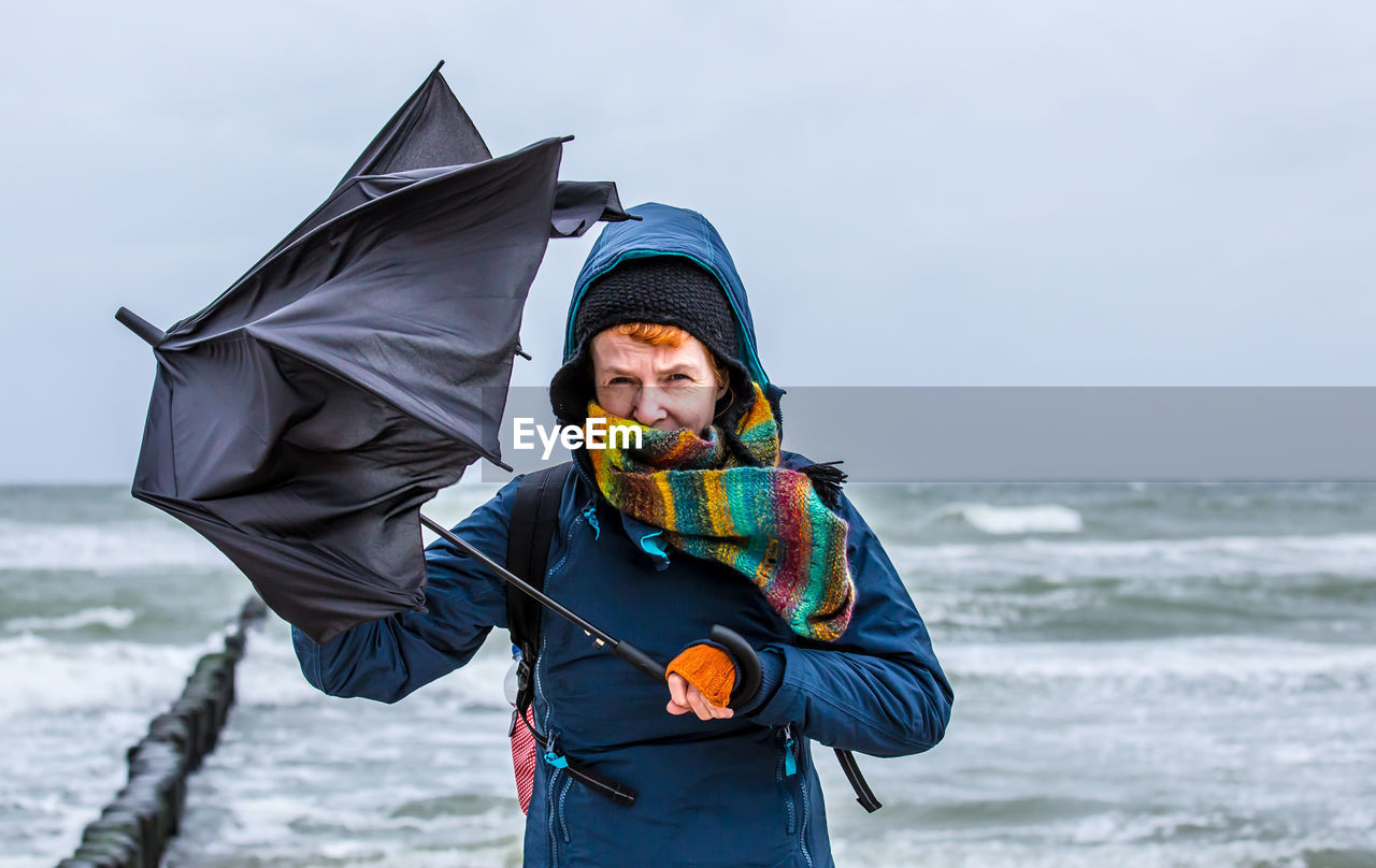 Portrait of woman holding damaged umbrella while standing at shore of beach against sky