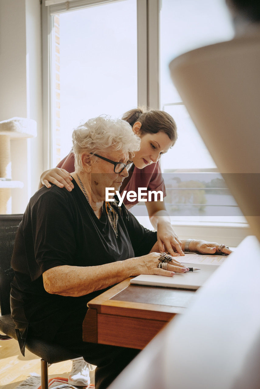 Female caregiver with hand on shoulder of senior woman sitting at desk in apartment