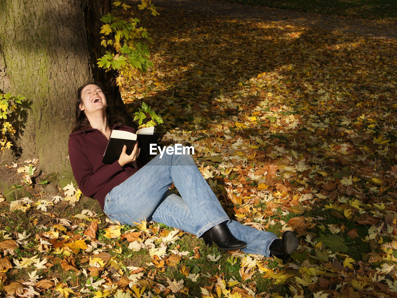 Full length of woman laughing while holding book by tree at park during autumn