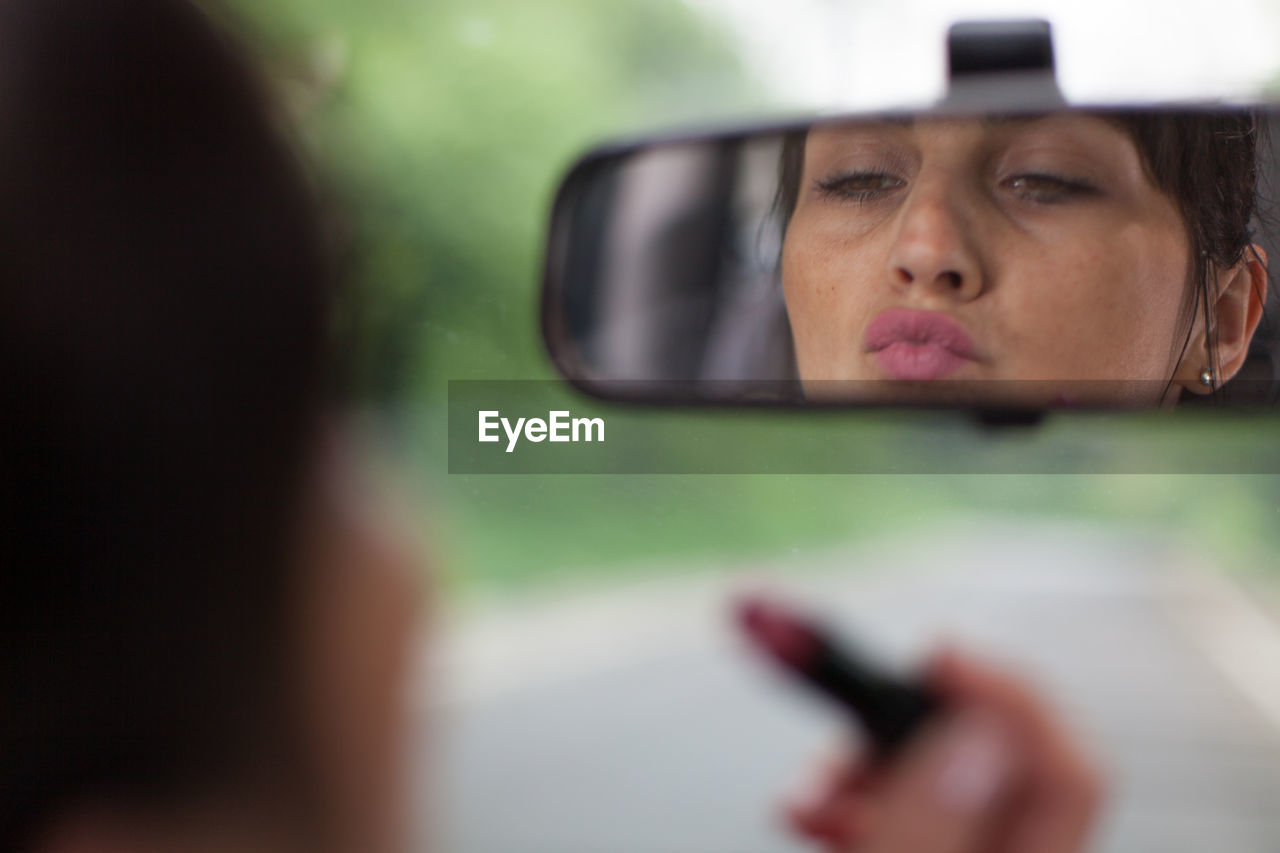 Close-up of mature woman applying lipstick reflecting on rear-view mirror