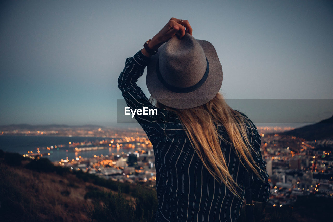 Rear view of woman by illuminated cityscape against clear sky during dusk