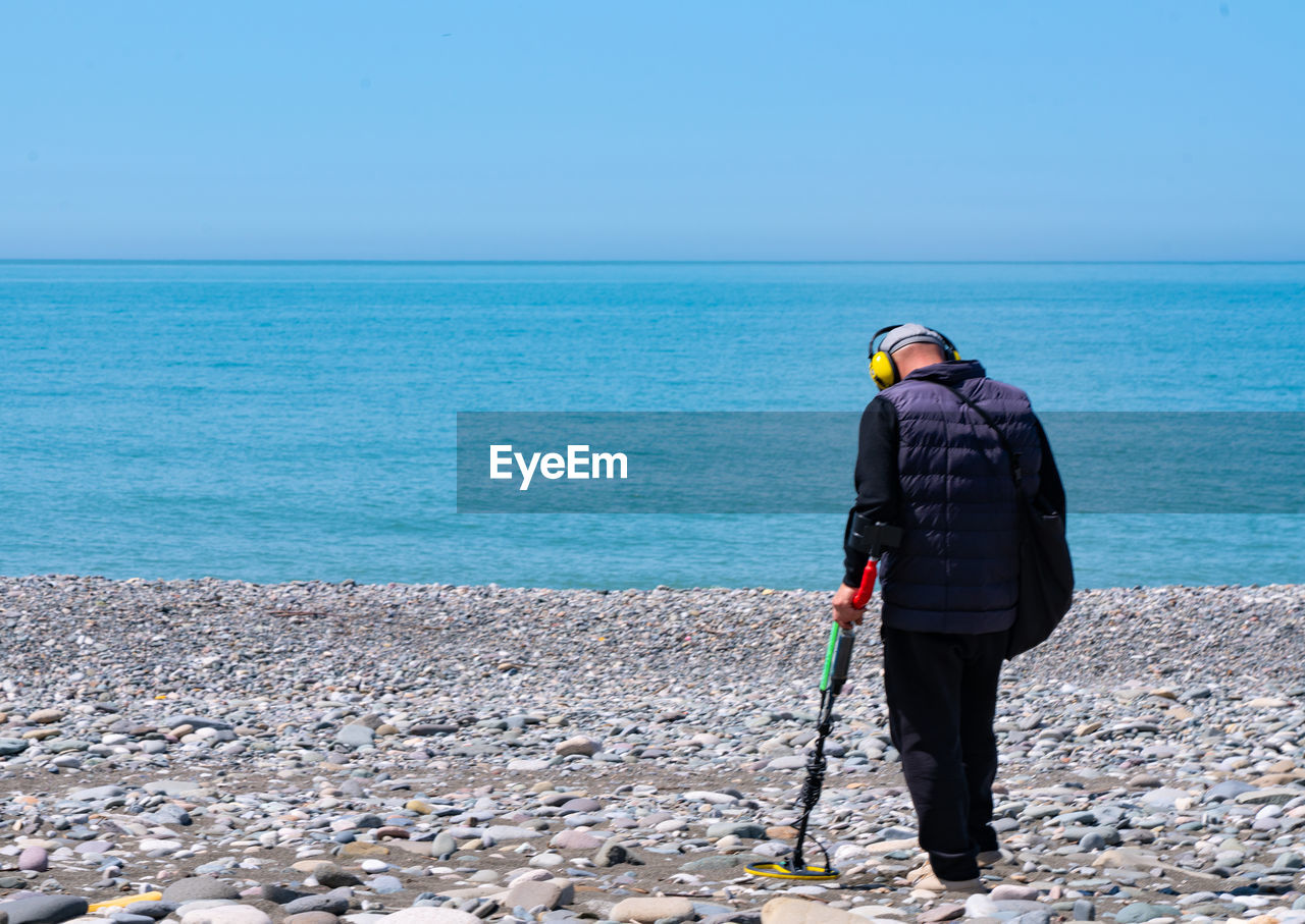 REAR VIEW OF MAN STANDING ON SHORE AGAINST SEA