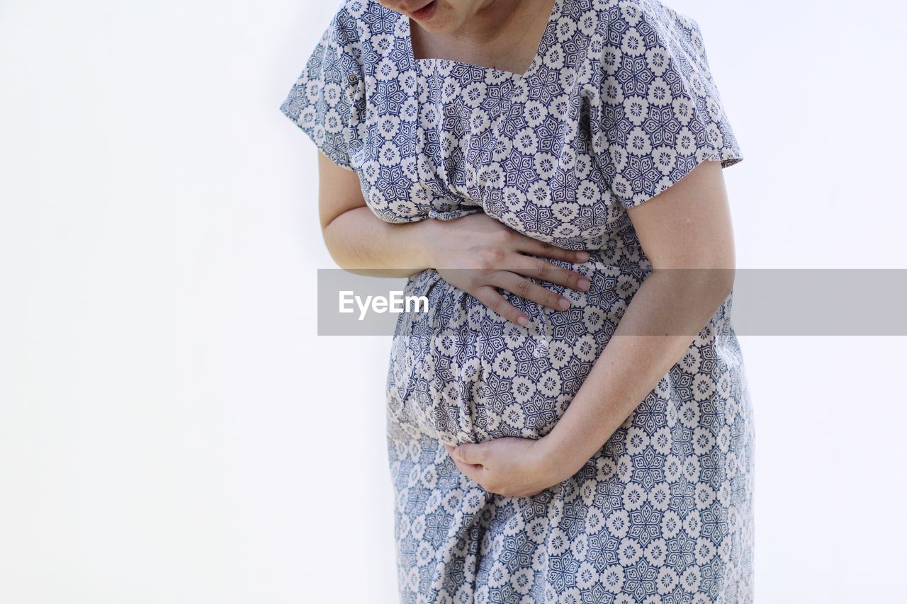 Midsection of pregnant woman touching abdomen while standing against white background