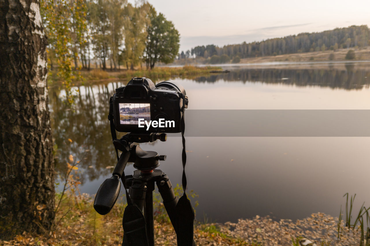 Black digital camera on tripod shooting foggy morning landscape at autumn lake with selective focus.