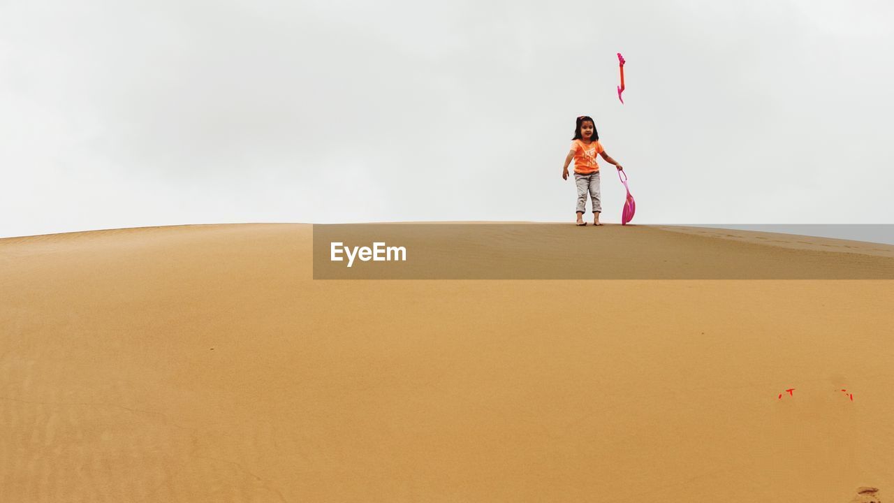 Full length of girl throwing shovel while standing on sand dune against sky