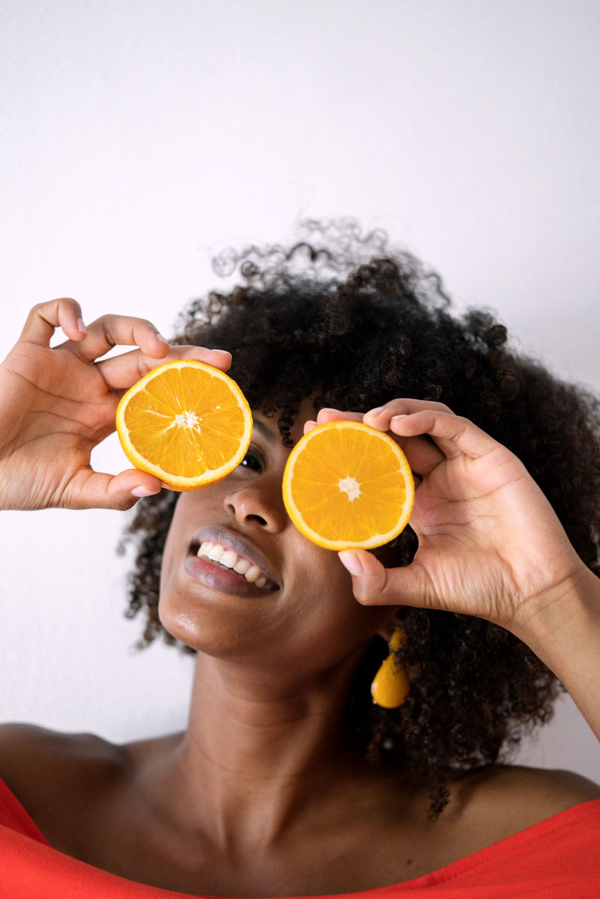 Black woman with afro hair holding cut orange halves in front of her eyes having fun