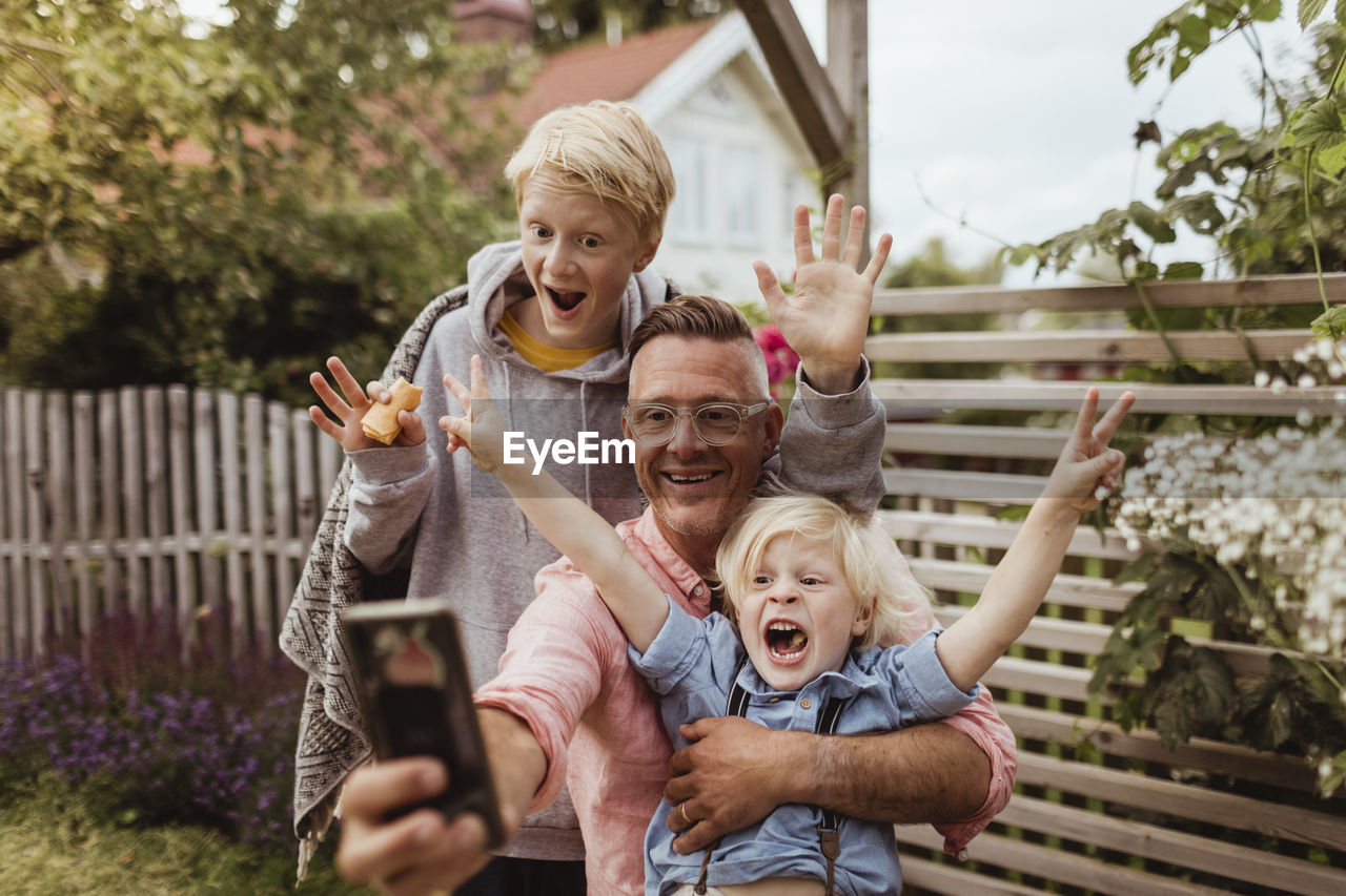 Cheerful father taking selfie with sons on smart phone in front yard at evening