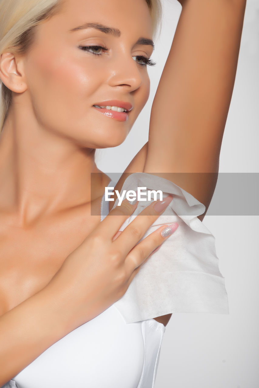 Smiling young woman cleaning armpit with tissue paper against white background