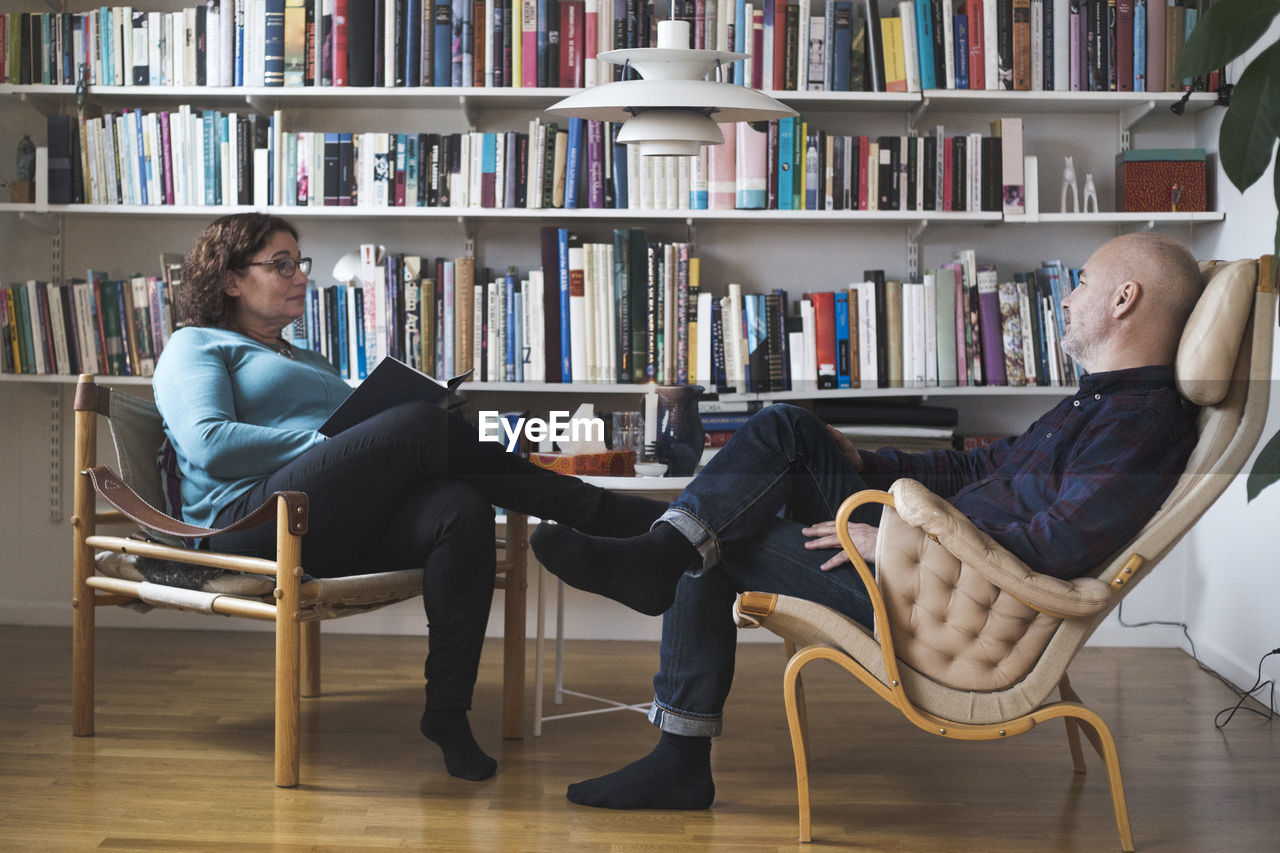 Mature female therapist talking to patient by bookshelf at home office