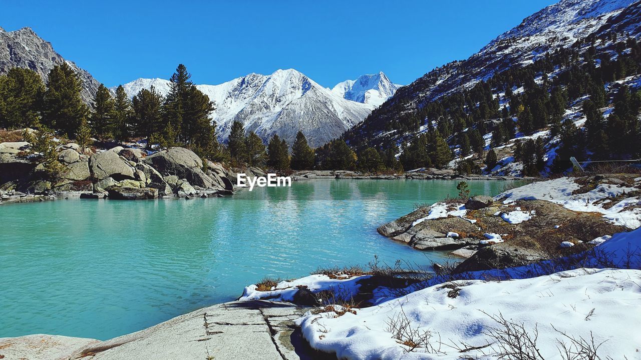 mountain, snow, scenics - nature, water, beauty in nature, cold temperature, winter, nature, mountain range, environment, tree, sky, lake, tranquil scene, landscape, tranquility, snowcapped mountain, pine tree, pinaceae, wilderness, land, plant, coniferous tree, day, travel destinations, blue, no people, pine woodland, travel, clear sky, non-urban scene, forest, glacial lake, sunlight, outdoors, mountain peak, idyllic, rock, sunny, leisure activity, tourism, activity, ice, adventure, frozen