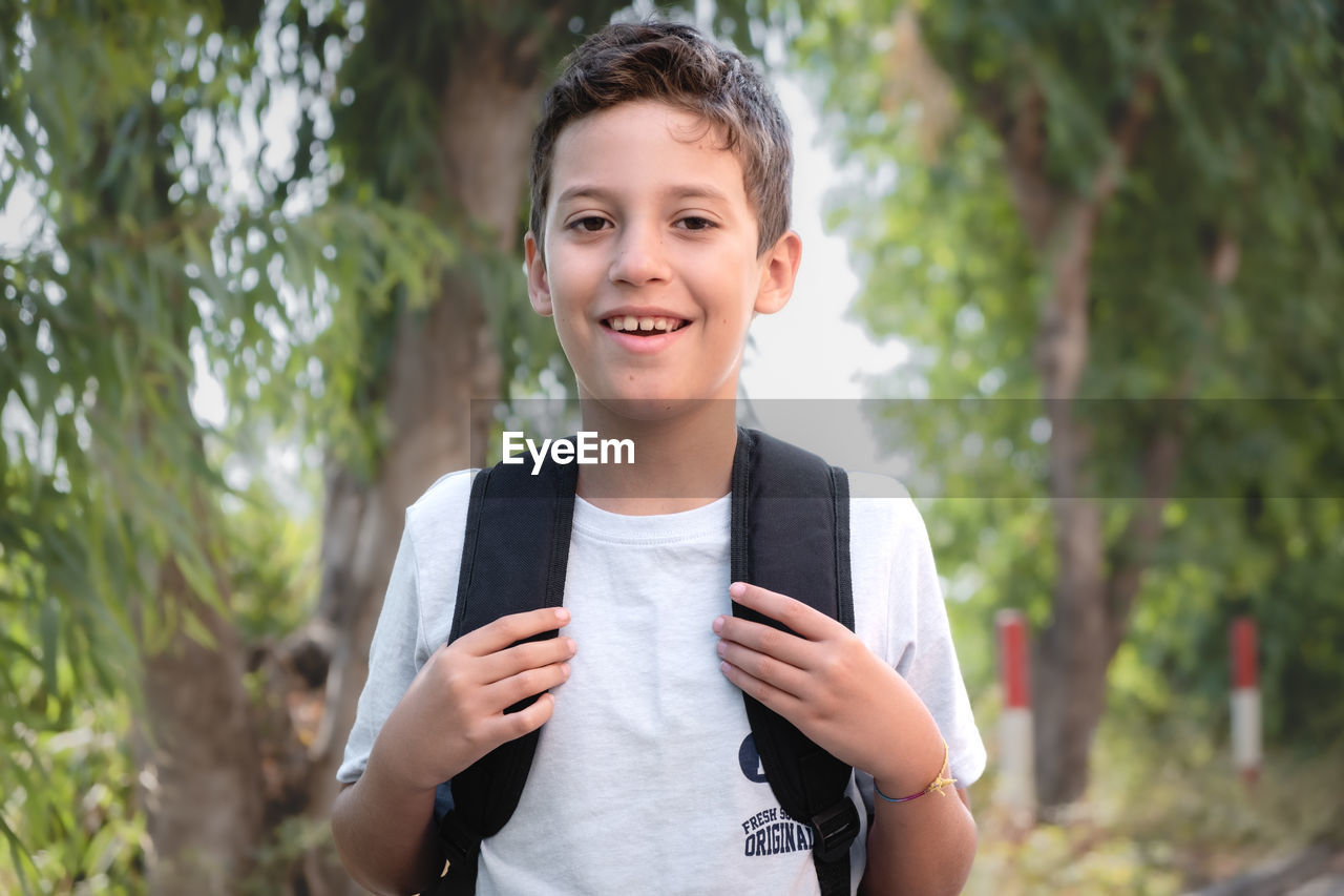 Portrait of a boy walking under the trees with a backpack looking at the camera