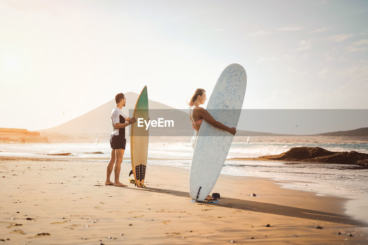 Man and woman standing with surfboard on beach against sea and sky