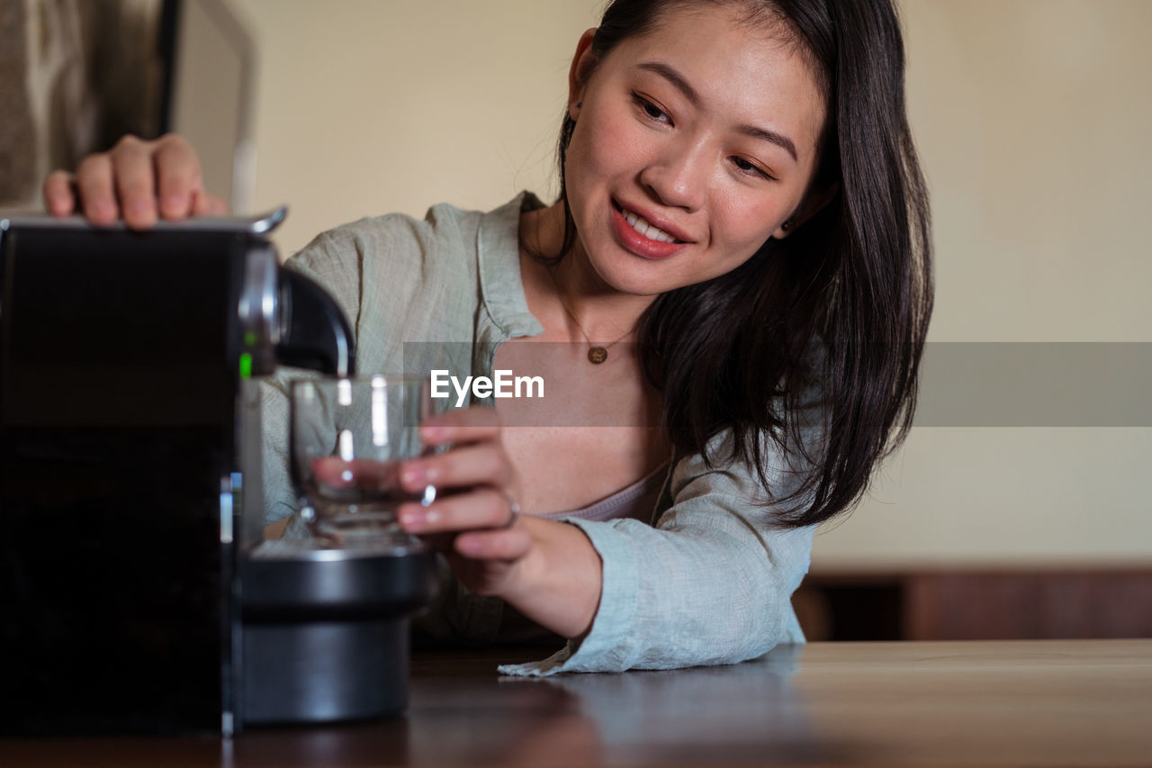 Young ethnic female against pod coffee maker pouring hot beverage with foam into glass in house kitchen