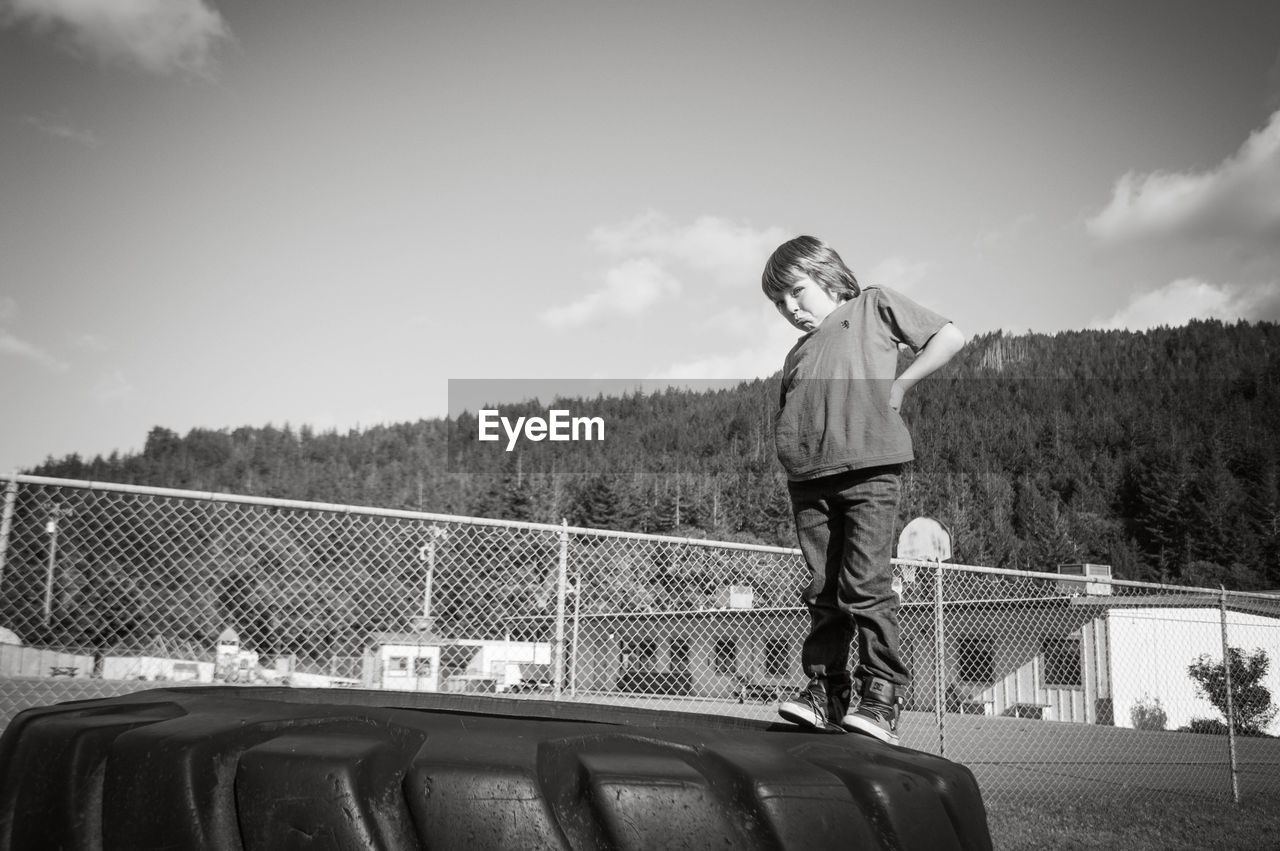 Portrait of boy making face while standing on tire against sky