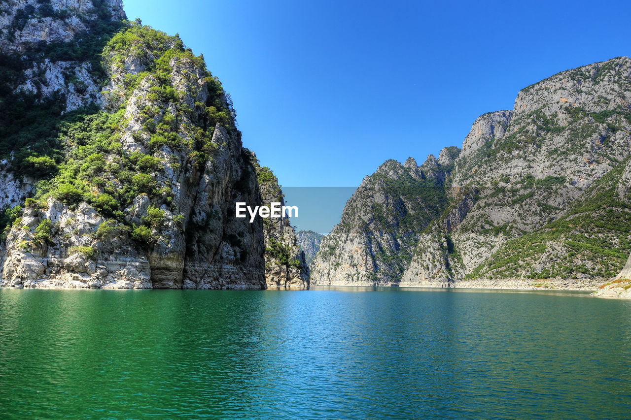 Scenic view of sea by mountains against clear blue sky