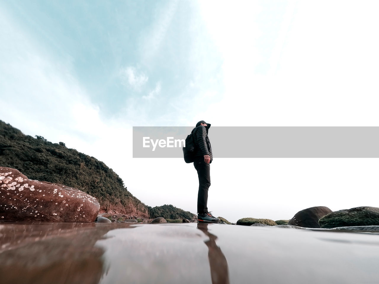 Water surface image of mature man standing on rock at beach against cloudy sky