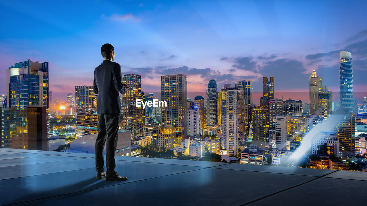 REAR VIEW OF MAN STANDING BY ILLUMINATED MODERN BUILDINGS AGAINST SKY