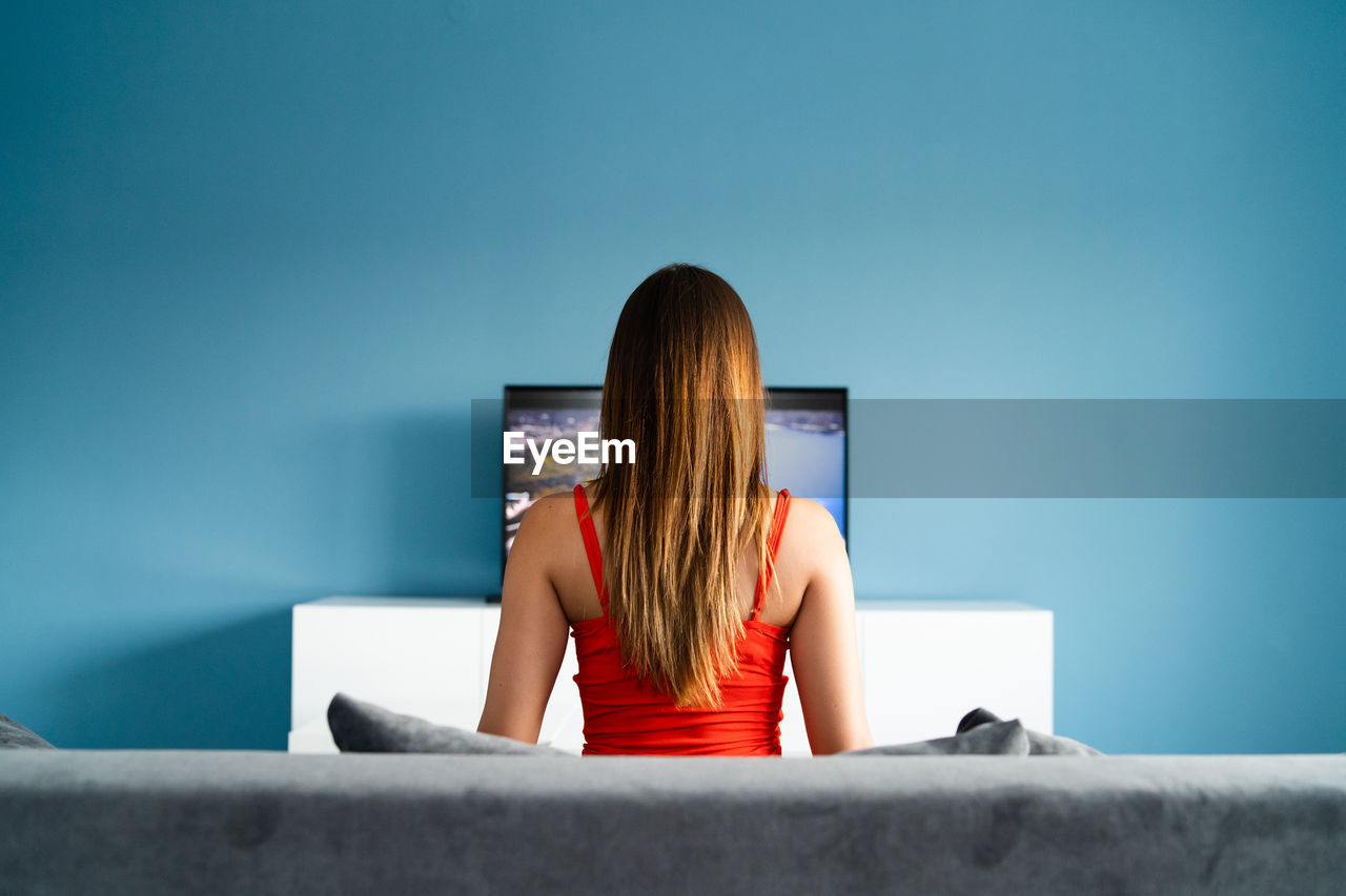 Rear view of woman looking at television set against blue wall at home