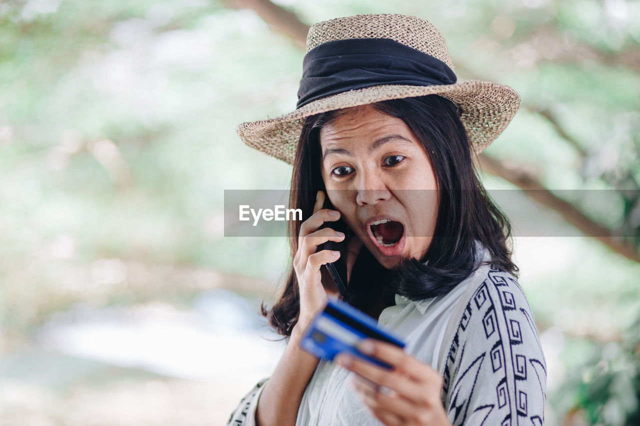 Mid adult woman with credit card talking over mobile phone while standing against trees