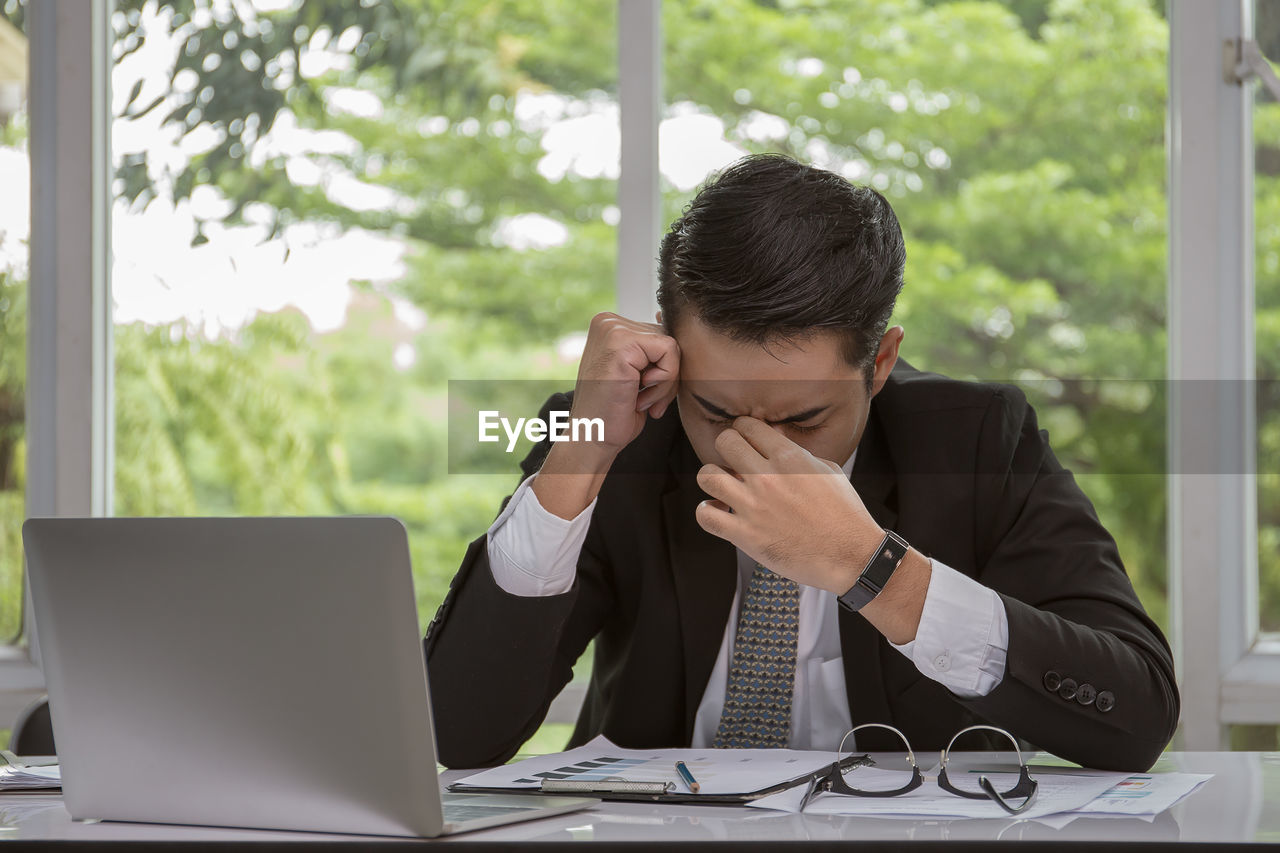 Tired businessman with laptop on desk sitting against window in office