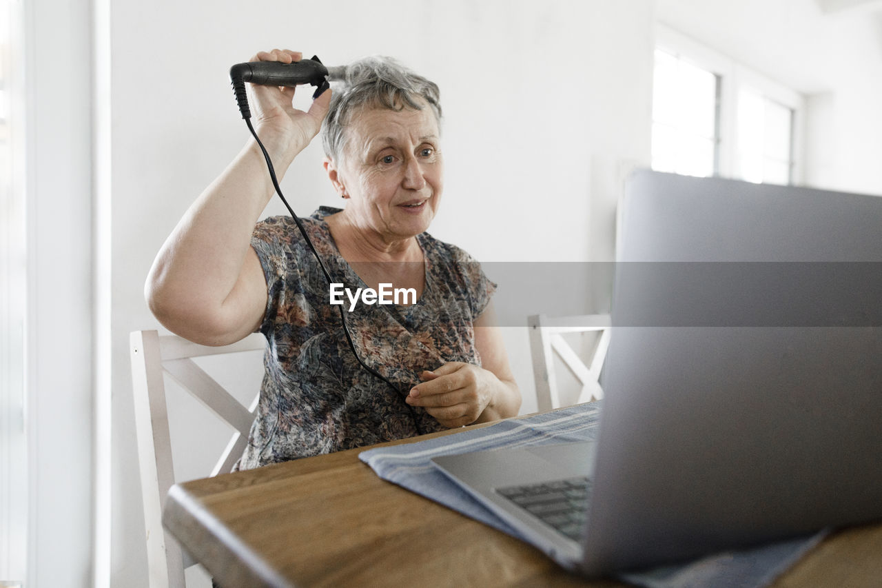 Senior woman sitting at table at home using laptop and doing her hair