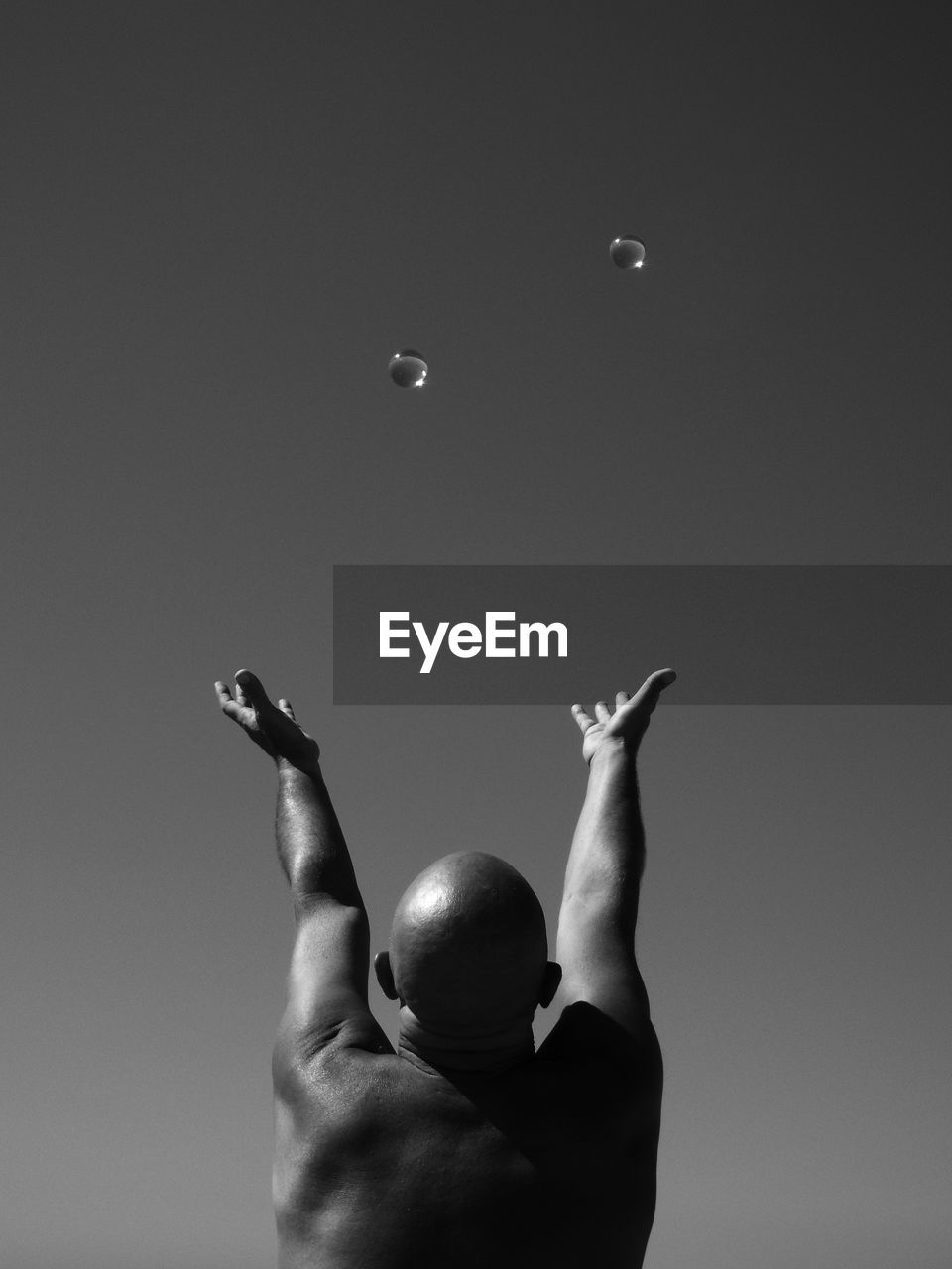 Rear view of bald man with arms raised towards bubbles against sky