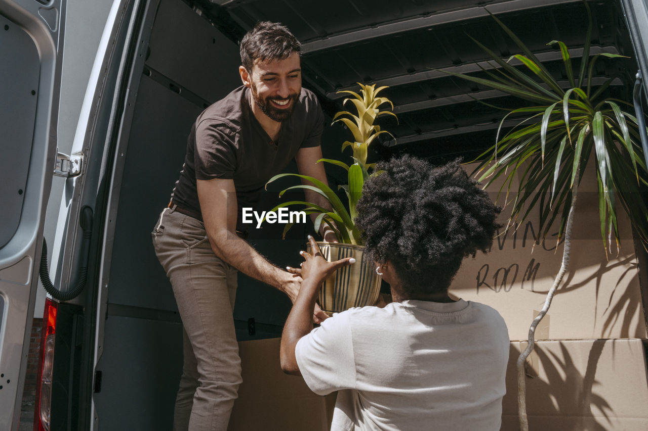 Smiling boyfriend passing plant to multiracial girlfriend near van trunk