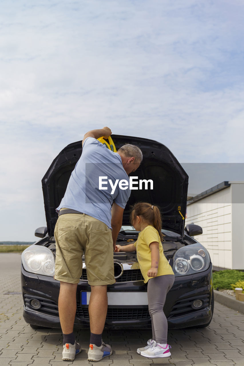 Girl helping mature man repairing car at parking lot