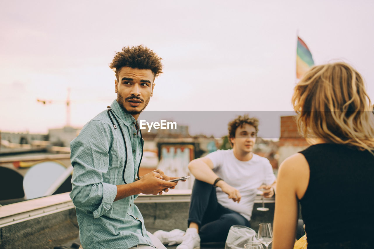 Young man text messaging on smart phone while sitting with friends on terrace at rooftop party