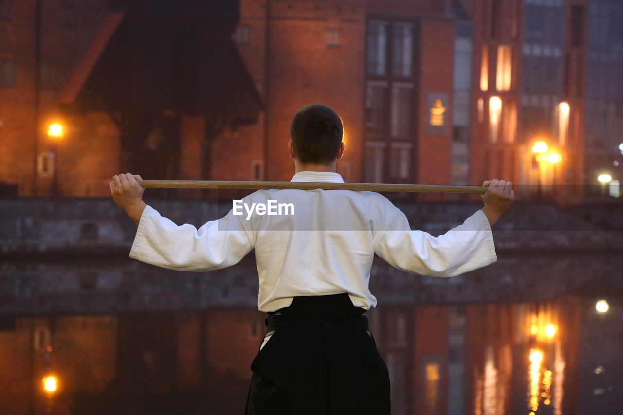Rear view of man practicing martial arts while standing against canal in city during sunset