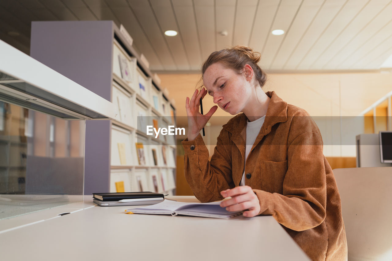 Young woman in casual clothes with hair bun touching head and writing in planner while sitting at table and studying in university library