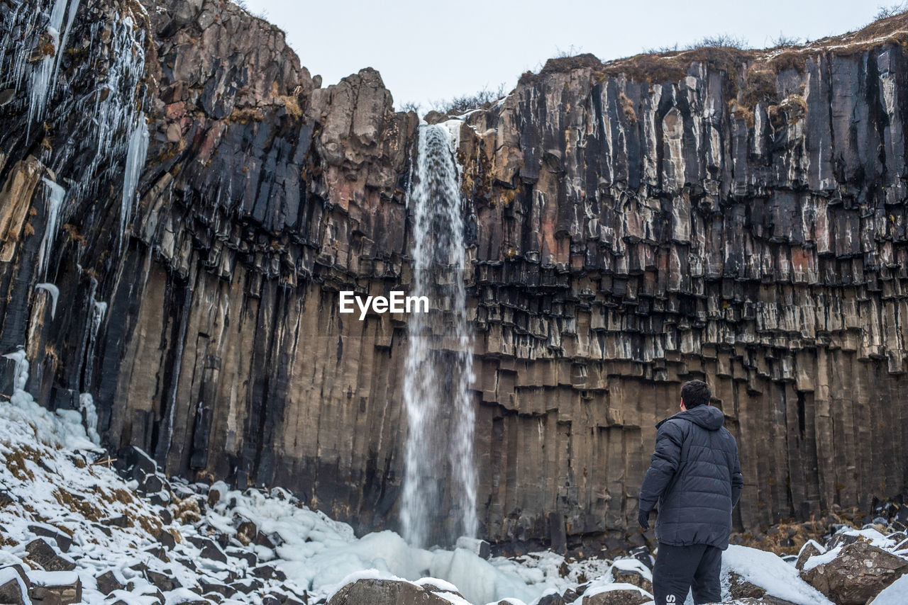 Rear view of man standing against waterfall and rock formation during winter