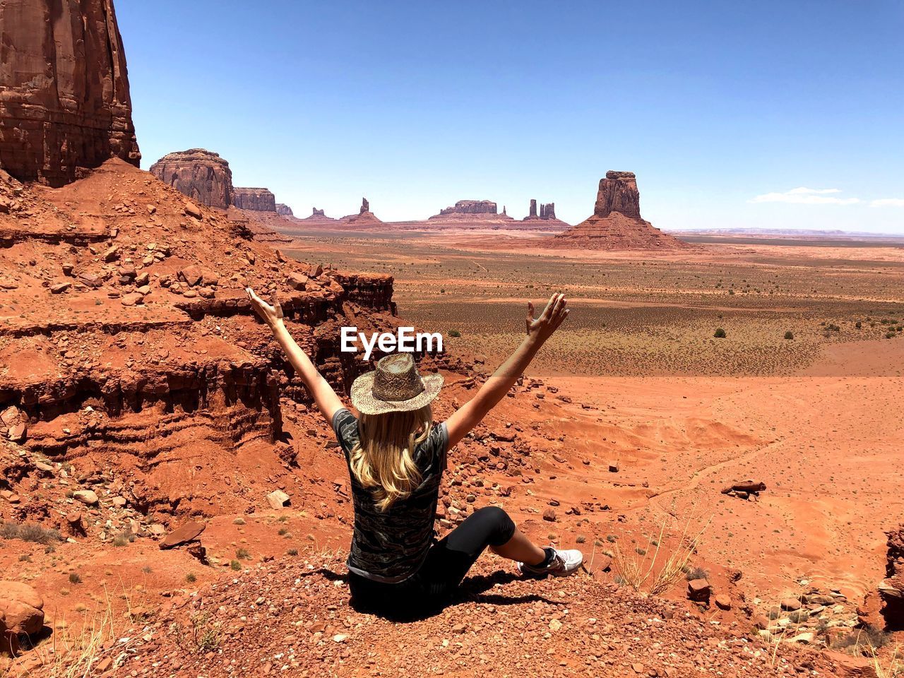 Woman with arms raised sitting on dirt road against blue sky