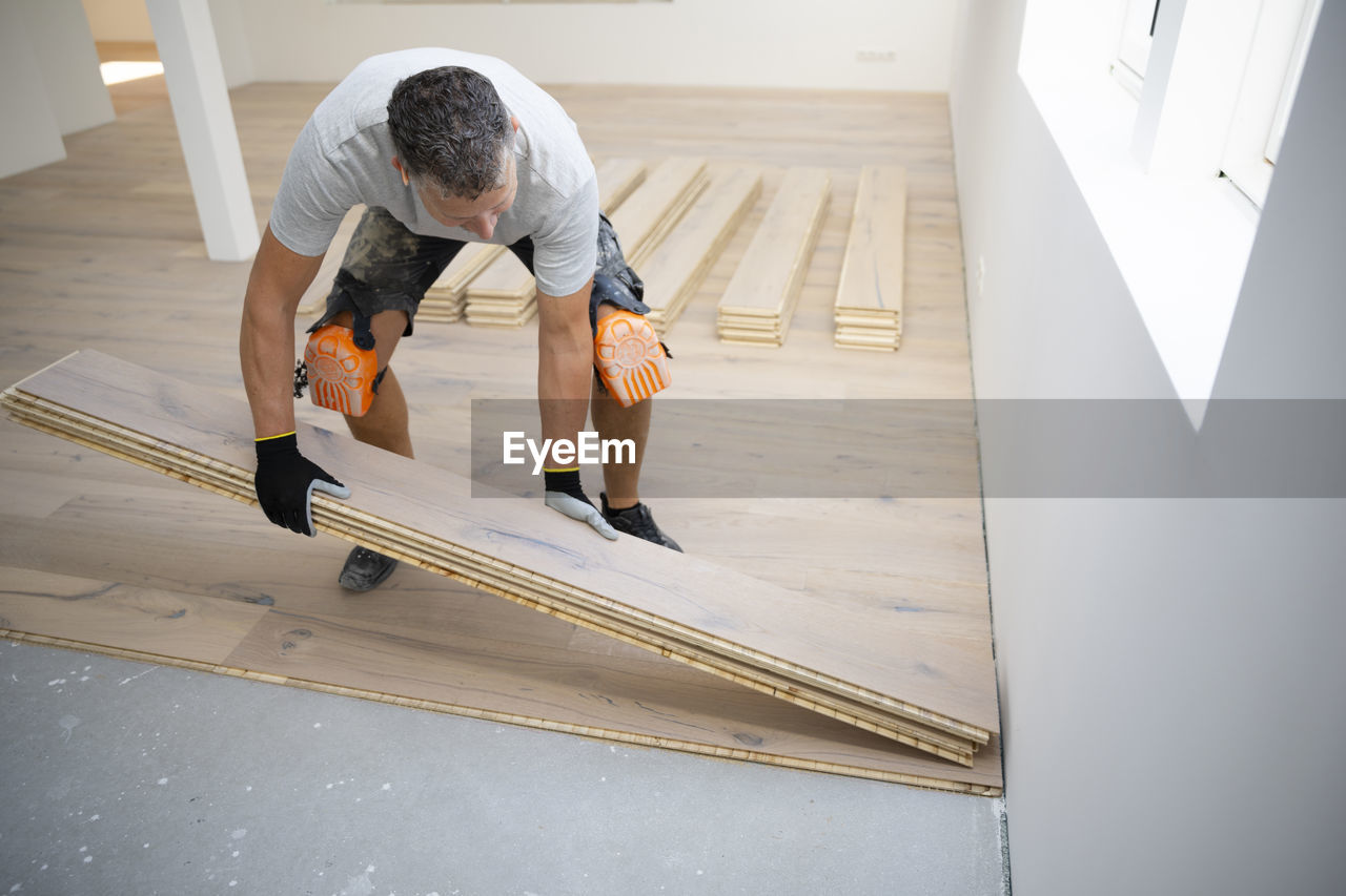 floor, adult, flooring, indoors, one person, men, wood, working, occupation, diy, home improvement, carpenter, business finance and industry, hardwood, improvement, skill, lifestyles, full length, looking down, concentration, person, construction industry, industry, home interior, accuracy, kneeling, craft, mature adult, business, craftsperson, looking, manual worker, carpentry, work tool, bending, building contractor, home ownership, casual clothing, room, wall, renovation, expertise