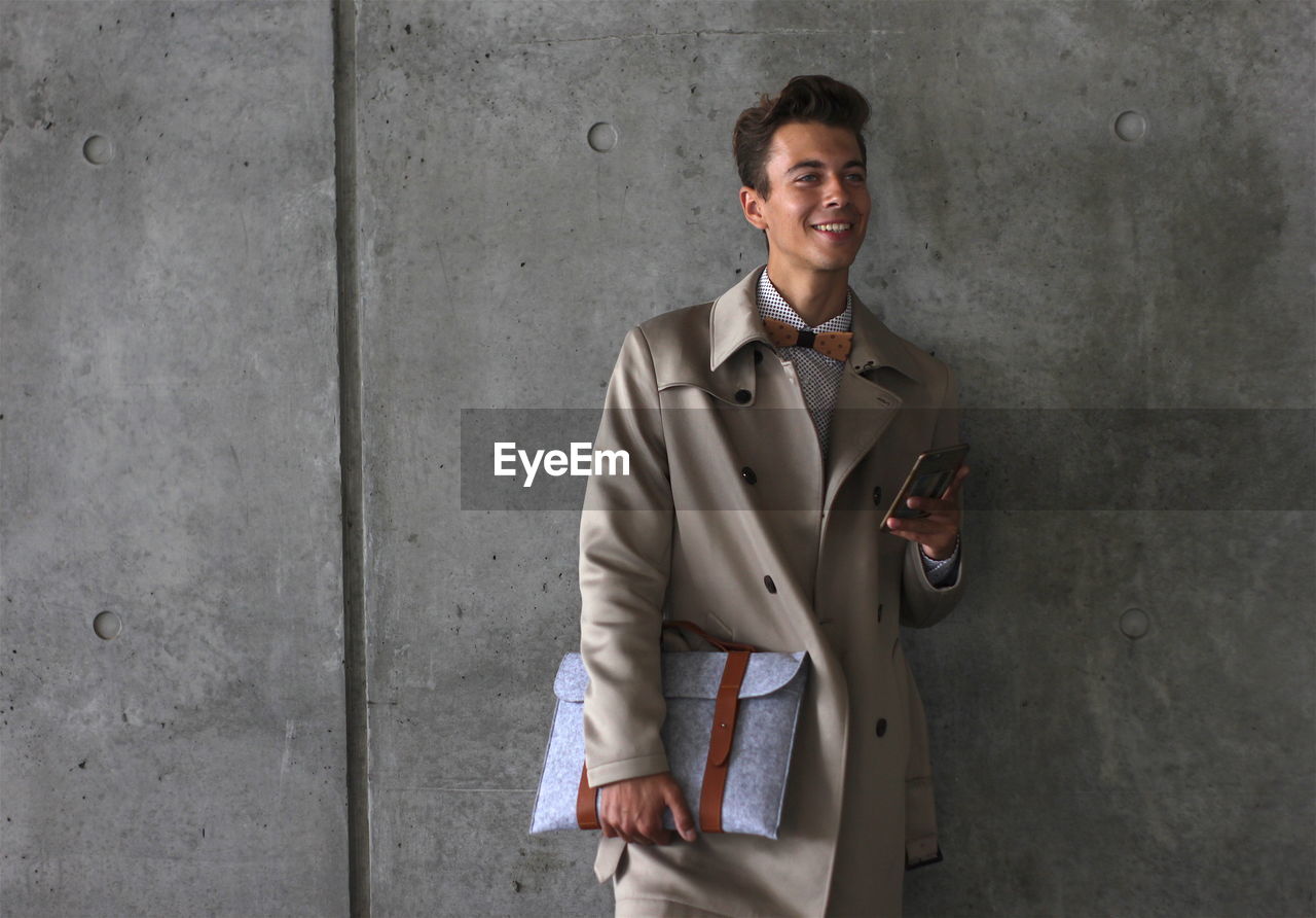 Young model holding mobile phone and bag while leaning on wall